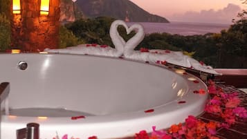 Una bañera blanca con pétalos de rosa y un arreglo con forma de corazón en el borde, rodeada de una vista panorámica de montañas y el océano.