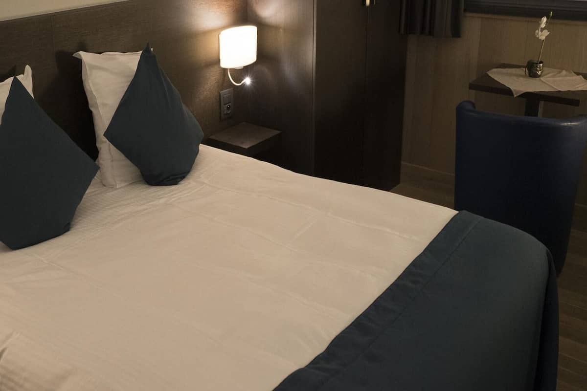 Ein Hotelzimmer mit Bett, Kissen, Nachttisch, Lampe und einem kleinen Tisch mit einer Vase.