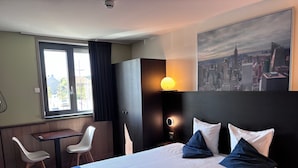 A hotel room with a bed, a desk, two chairs, a cityscape painting, and a window with curtains. - Hotel Orchidee (Aalter)