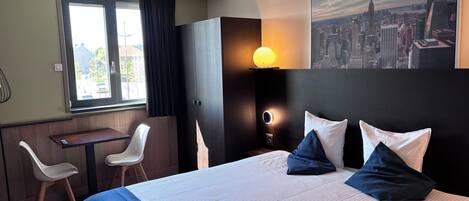 Una habitación de hotel con cama, escritorio, dos sillas, un cuadro de un paisaje urbano y una ventana con cortinas.