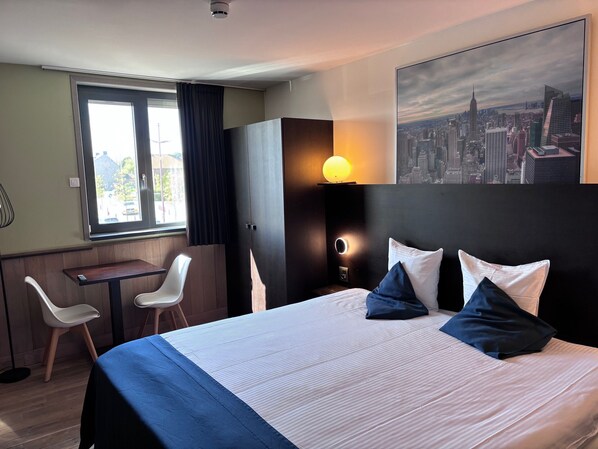 Una habitación de hotel con cama, escritorio, dos sillas, un cuadro de un paisaje urbano y una ventana con cortinas.