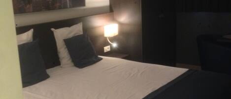 A hotel room with a large bed, a nightstand with a lamp, a painting on the wall, and a dark-colored headboard.