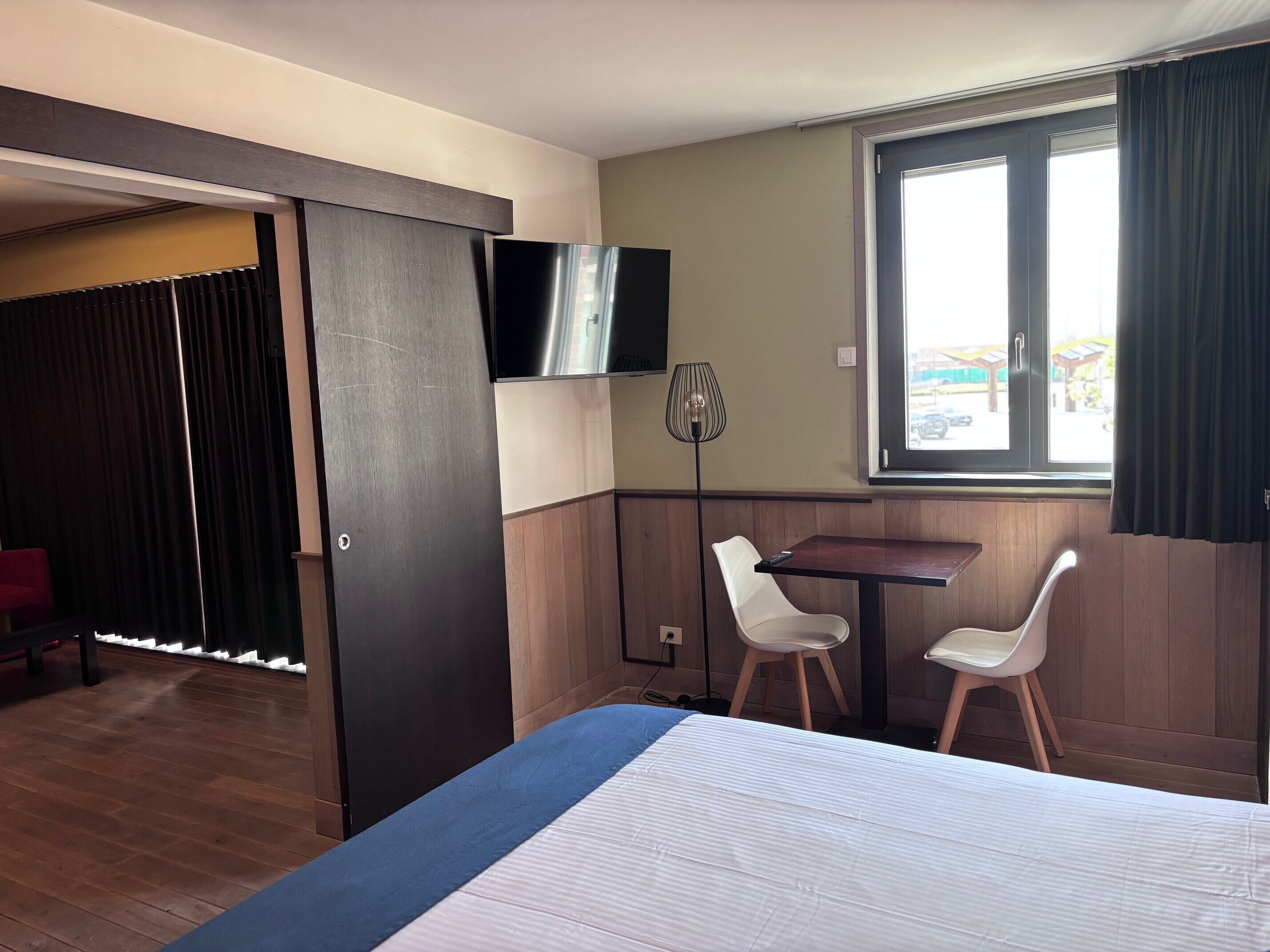Quarto de hotel com cama, duas cadeiras, mesa, televisão e janela com cortinas.