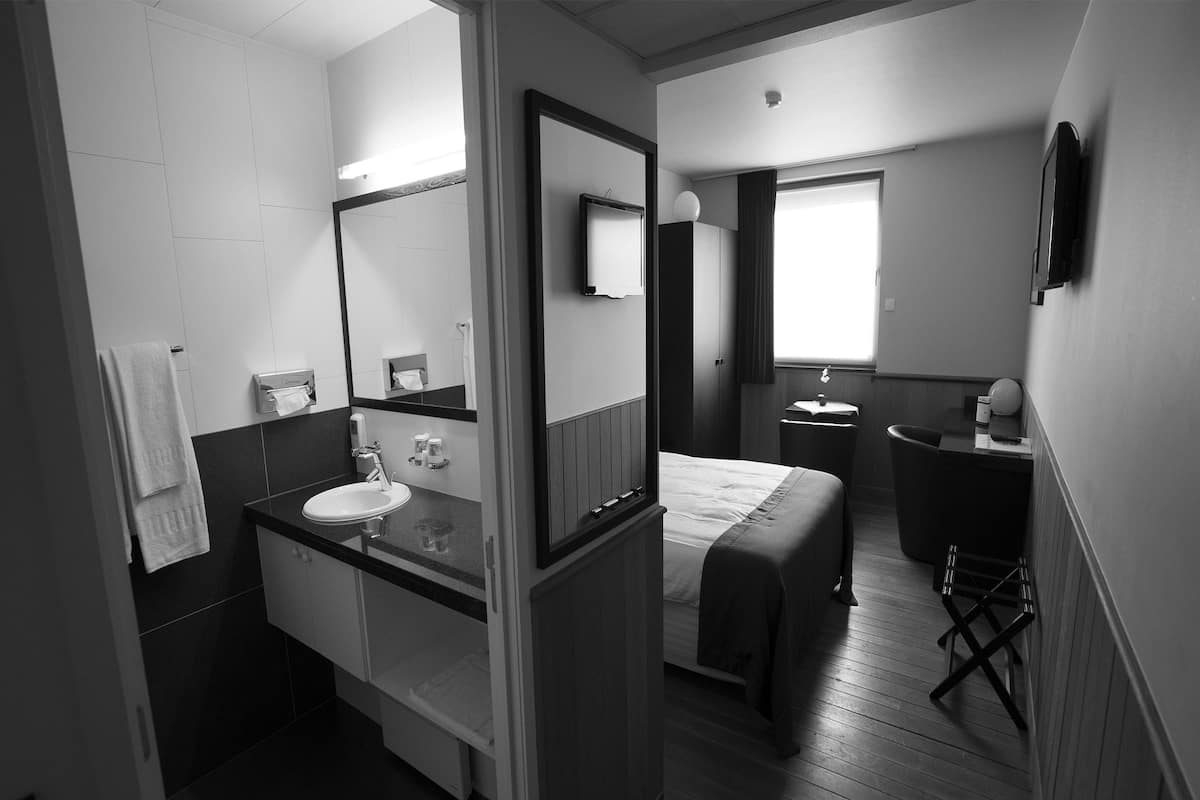 Ein kleines Hotelzimmer mit Bad, ausgestattet mit Waschbecken, Spiegel und Handtuchhalter. Ein Bett mit Decke steht an der Wand, und ein kleiner Tisch mit Stuhl befindet sich in der Nähe des Fensters.