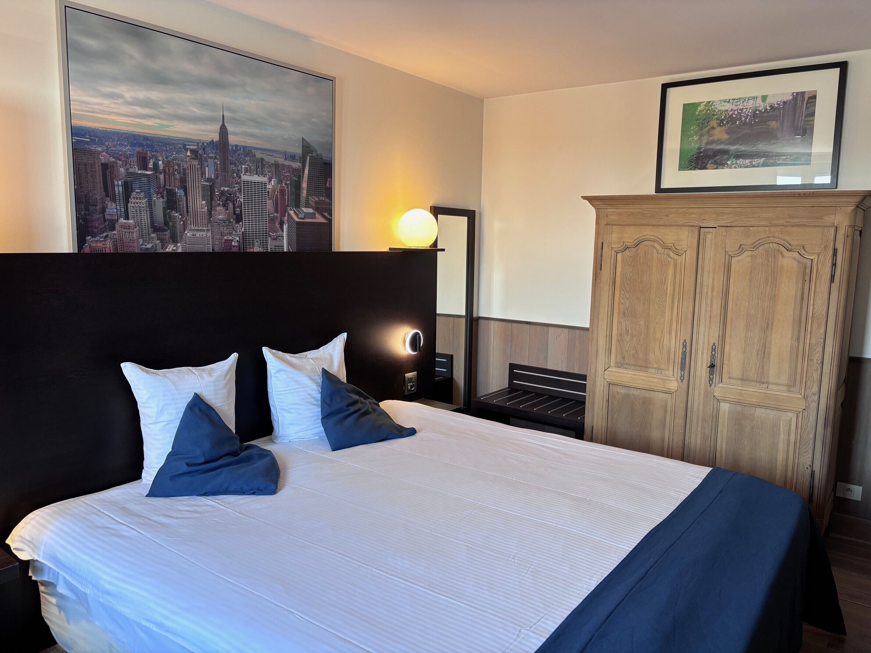 Une chambre d’hôtel avec un grand lit, un tableau représentant un paysage urbain, une armoire en bois et une table de chevet avec une lampe.