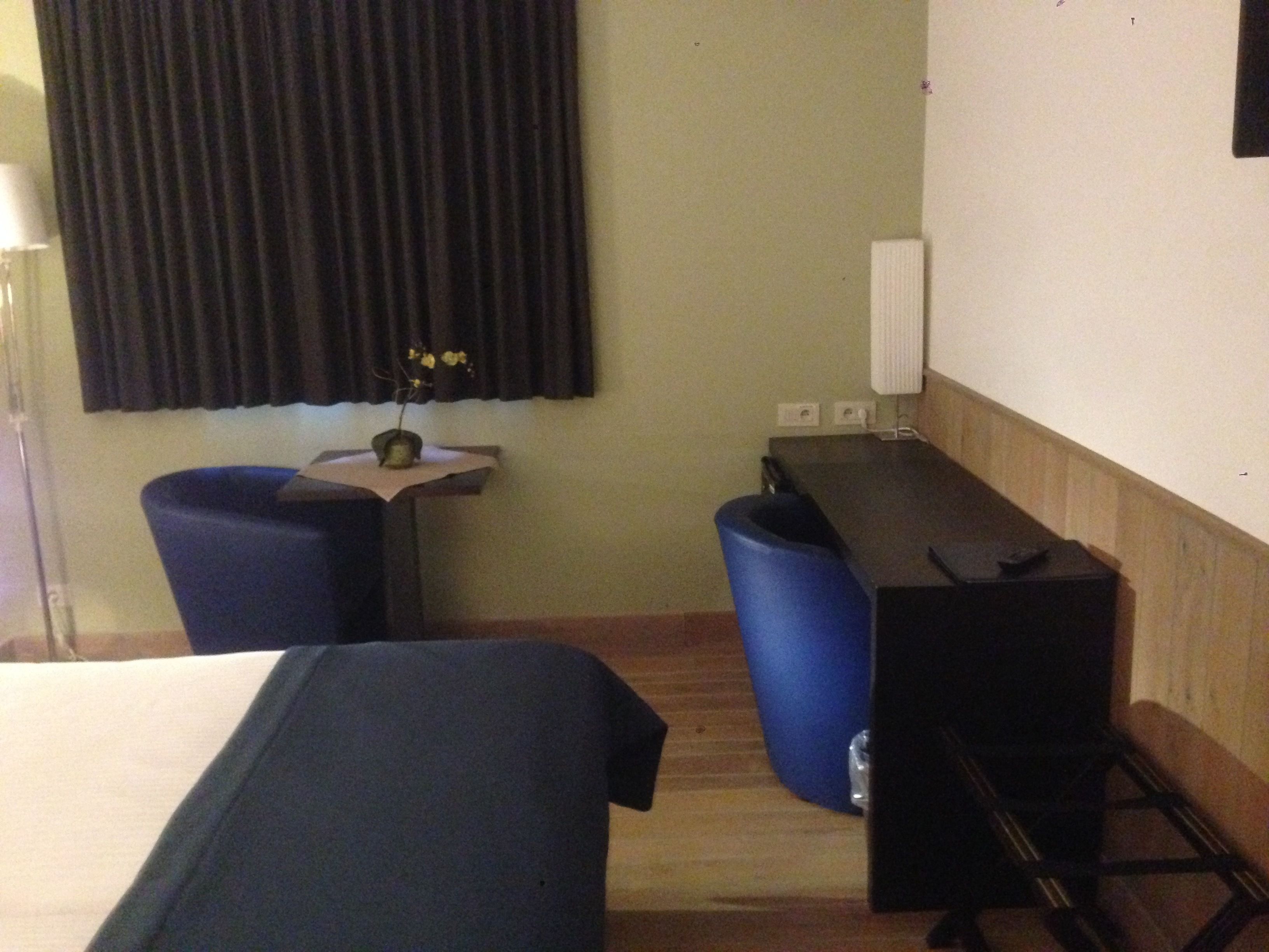 A hotel room with two beds, a desk, a chair, a small table with a vase, and a radiator.