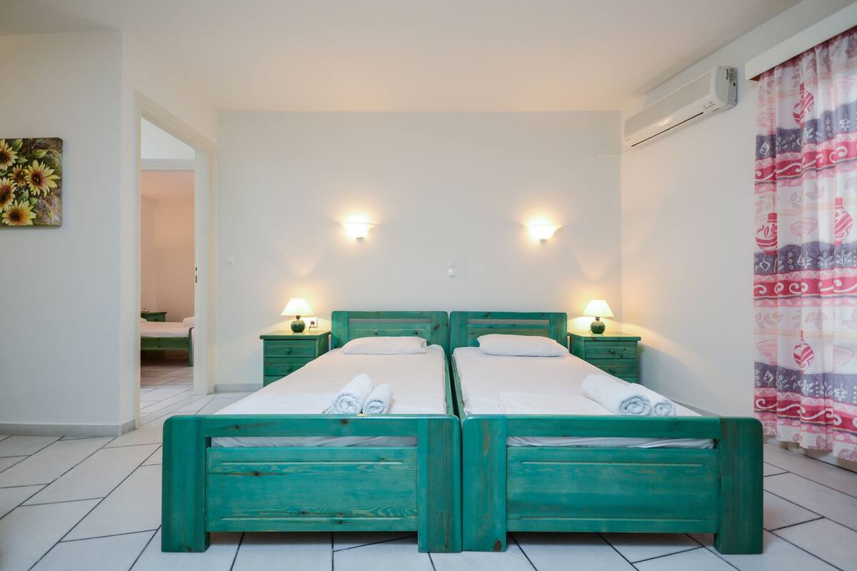 A room with two twin beds, each with a nightstand and a lamp. There is a painting on the wall and a patterned curtain on the window.