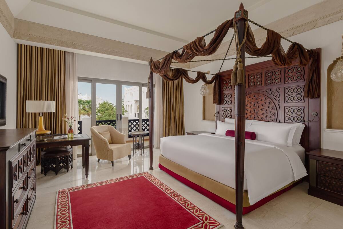 Um quarto espaçoso com uma cama de dossel, um espaço de estar com uma cadeira e uma mesa, e uma varanda com vista para a cidade.