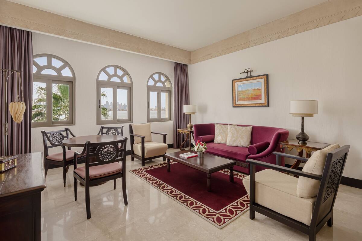 Uma sala de estar espaçosa com um sofá vermelho, poltronas bege, uma mesa de centro de madeira e um tapete estampado. Janelas amplas com arcos proporcionam vistas da cidade.