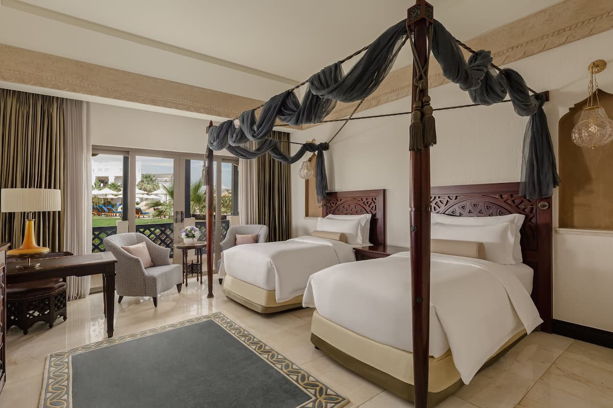 Quarto de hotel com duas camas, uma escrivaninha, uma cadeira e vista para o exterior.