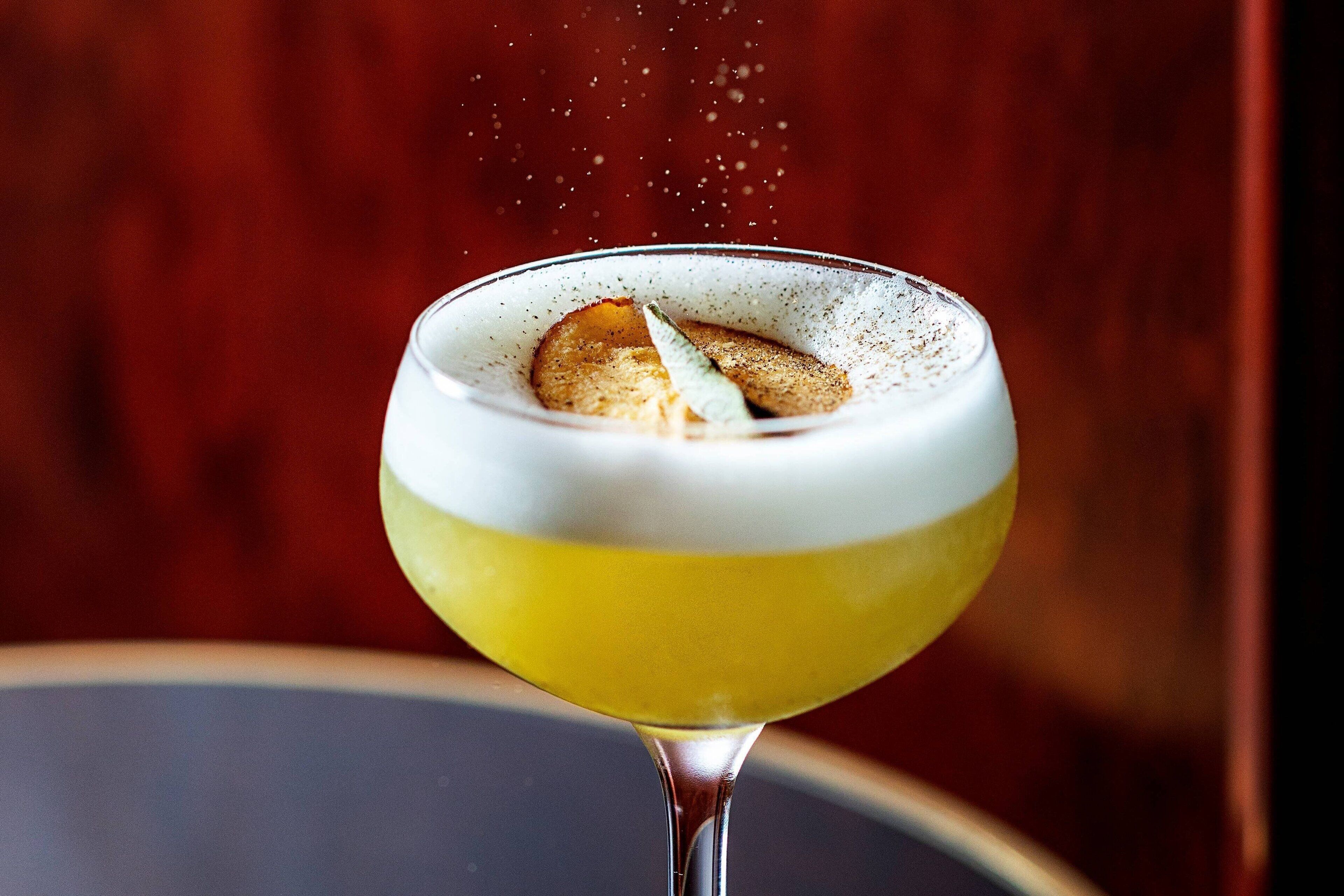 A cocktail with a frothy top, a slice of orange, and a sprig of mint, served in a stemmed glass.