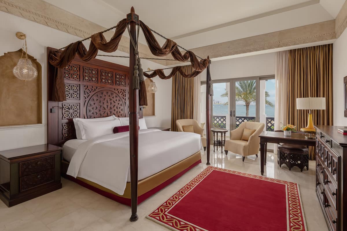Um quarto espaçoso com uma cama de dossel, um tapete vermelho, uma mesa de cabeceira de madeira, uma escrivaninha com uma luminária e vista para o mar.