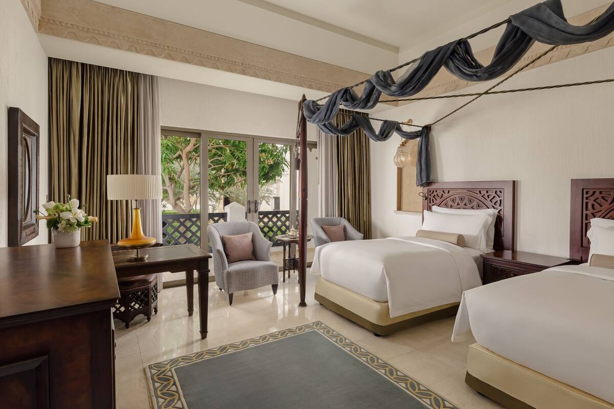 Quarto de hotel com duas camas, uma escrivaninha, uma cadeira e vista para o exterior.