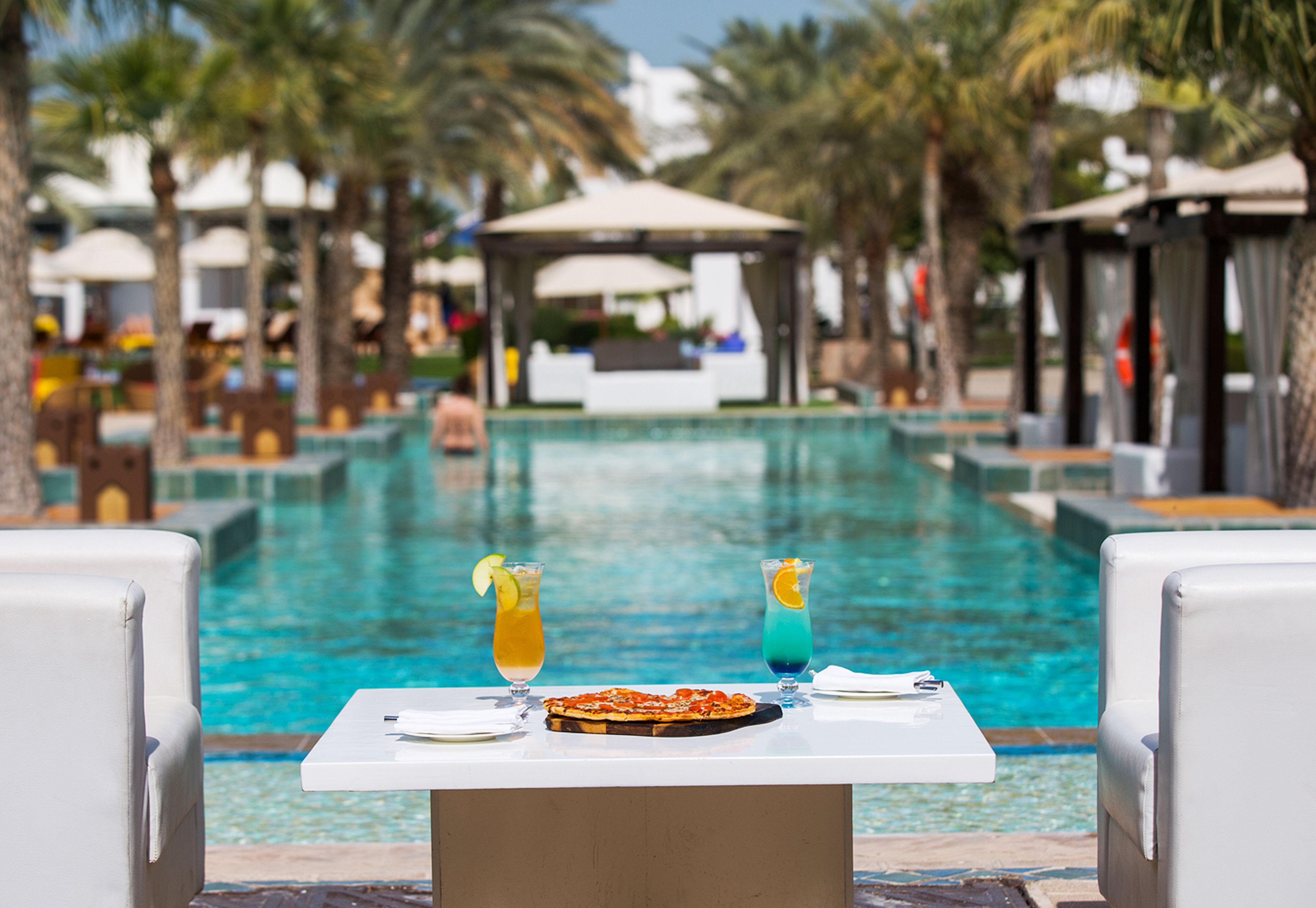 Un espace de restauration aménagé au bord de la piscine, avec une table dressée pour deux, donnant sur une piscine, entouré de palmiers et de chaises longues.