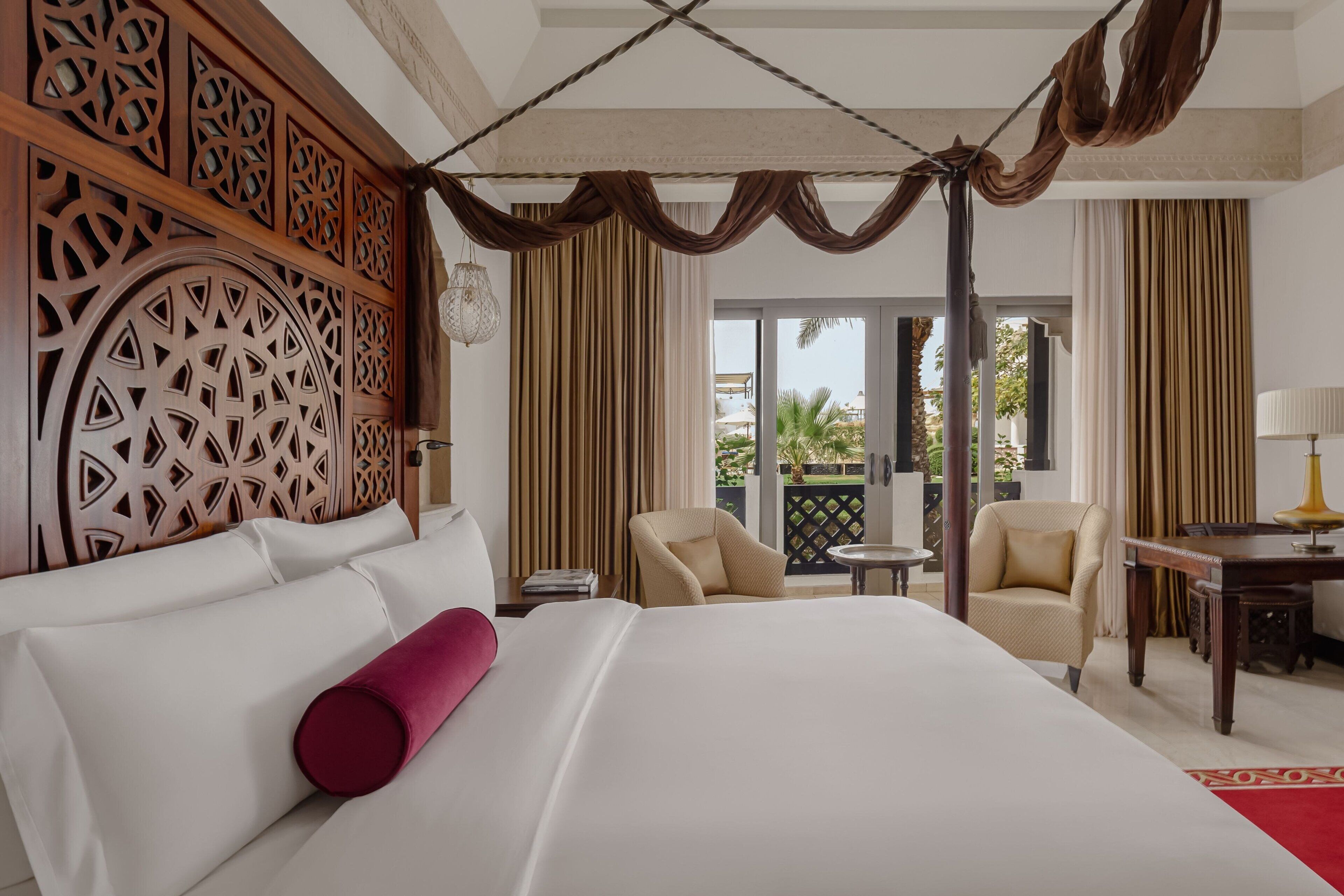 A hotel room with a large bed, two chairs, a small table, and a balcony with curtains.