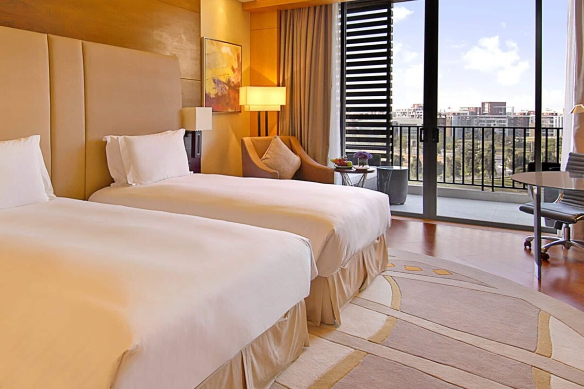 Habitación de hotel con dos camas, un escritorio, una silla, una lámpara y vistas a la ciudad a través de amplios ventanales.
