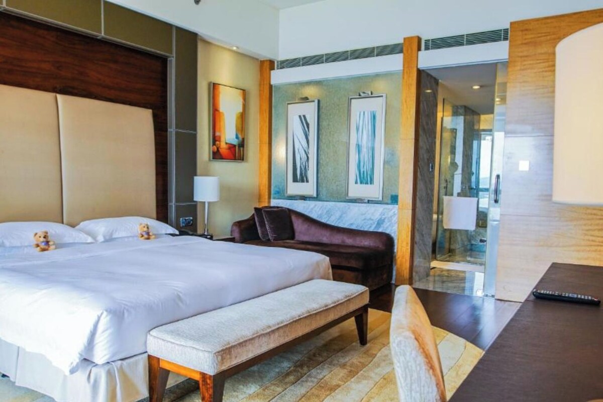 Habitación de hotel con una cama grande, un banco, un escritorio y una silla. Se observan cuadros en la pared y un cabecero de madera.