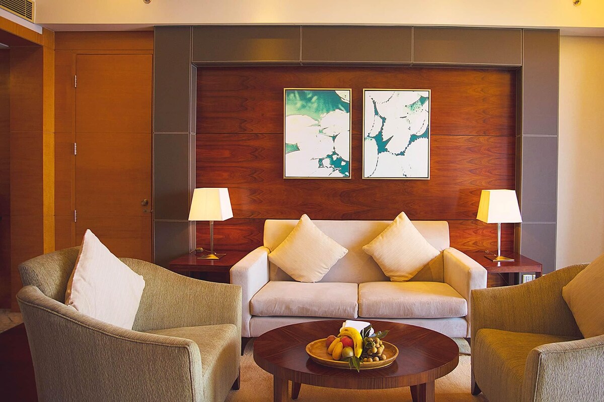 Habitación de hotel con sofá, dos sillones, una mesa de centro con fruta y dos lámparas de pared.