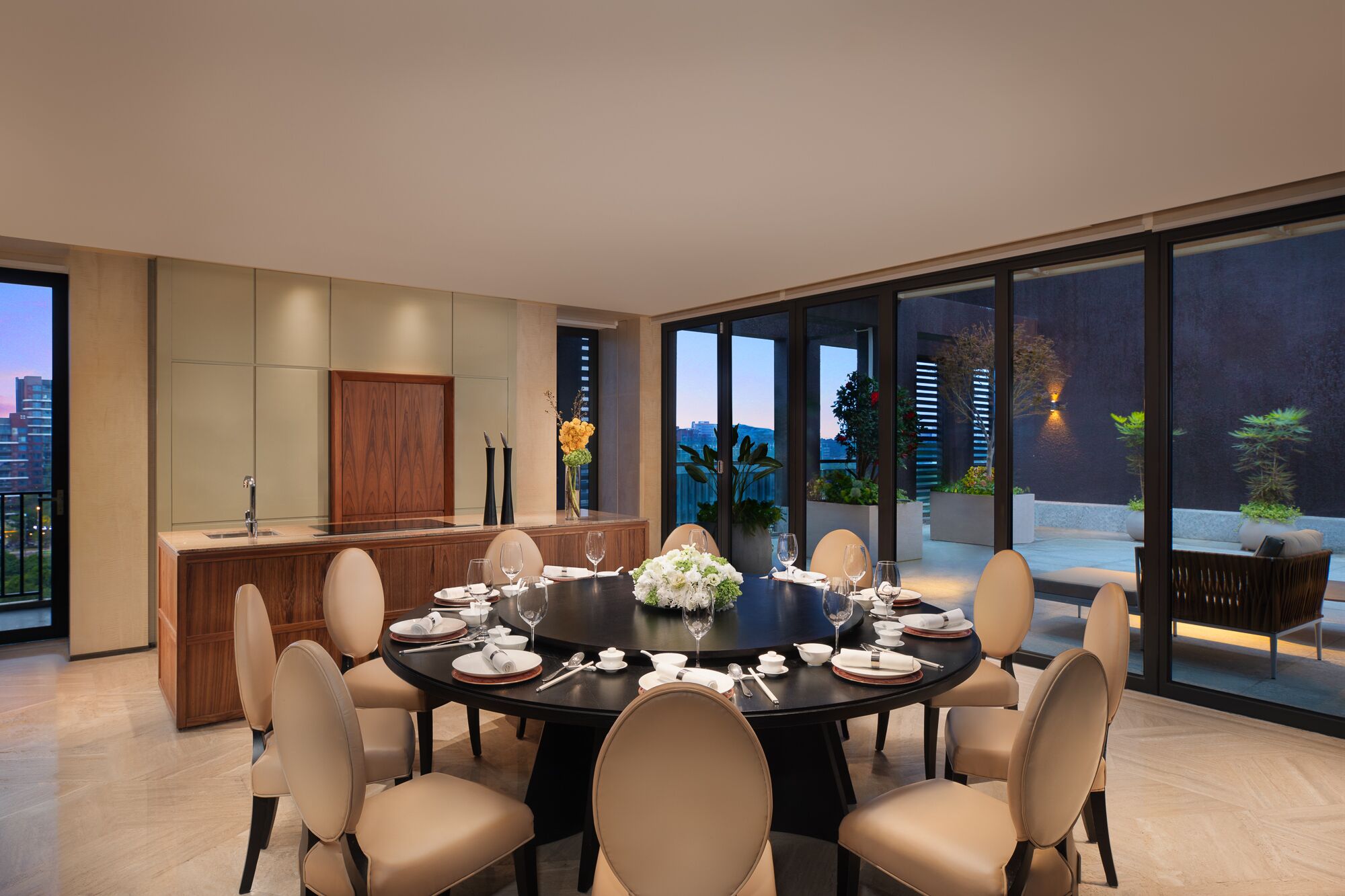 A modern dining room with a round table set for six, surrounded by beige chairs. A wooden cabinet and a large window with a city view are visible.