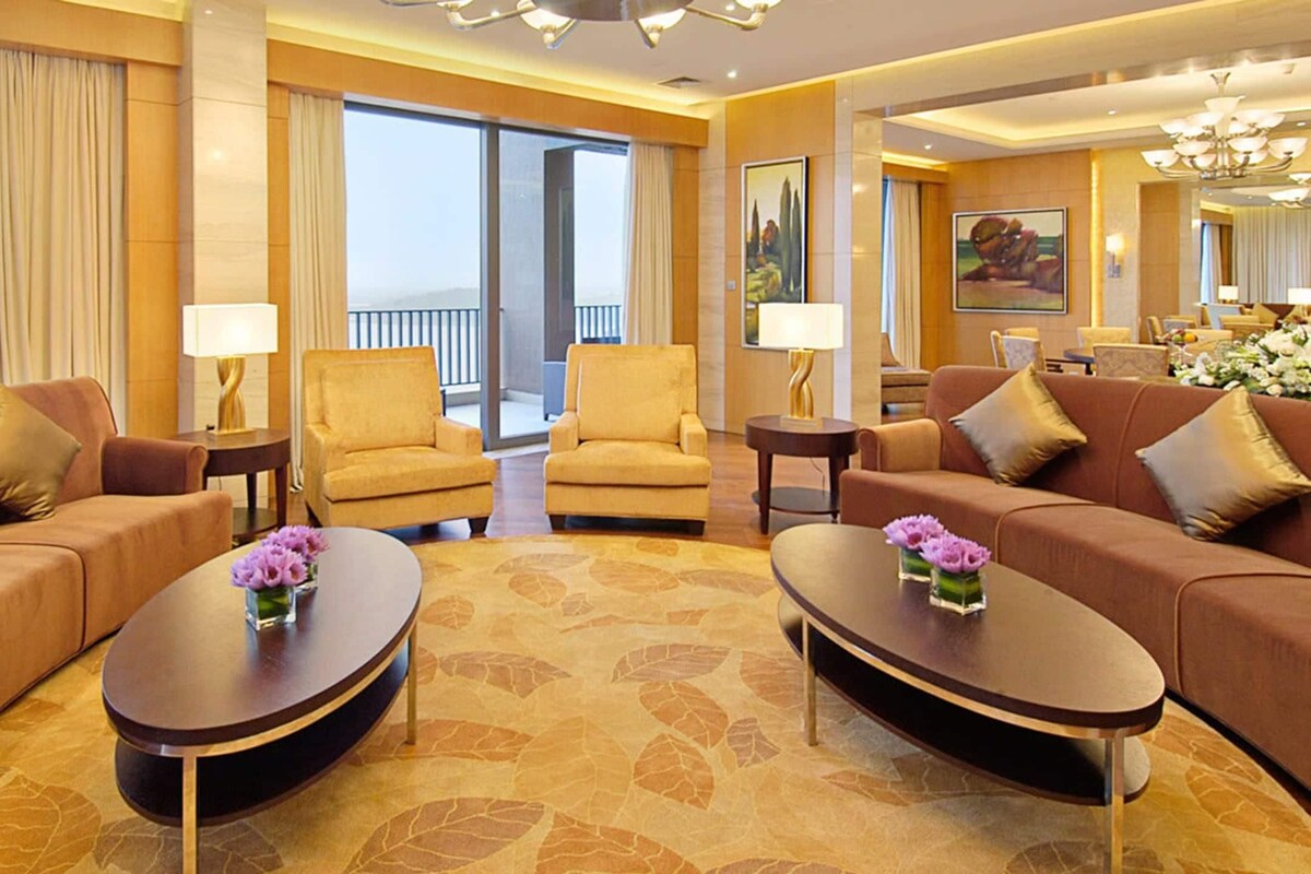 Habitación de hotel con un ventanal grande, dos sofás, dos sillones, una mesa de centro y una mesa de comedor.