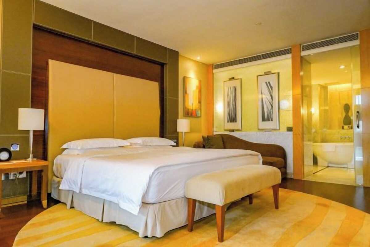 Habitación de hotel con una cama grande, mesitas de noche, un sofá y un baño visible a través de una puerta de cristal.