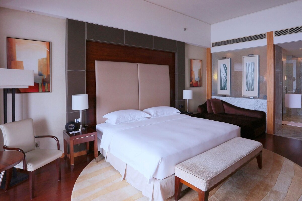 Habitación de hotel con una cama grande, mesitas de noche, una silla, un banco y un baño con cabina de ducha de vidrio.