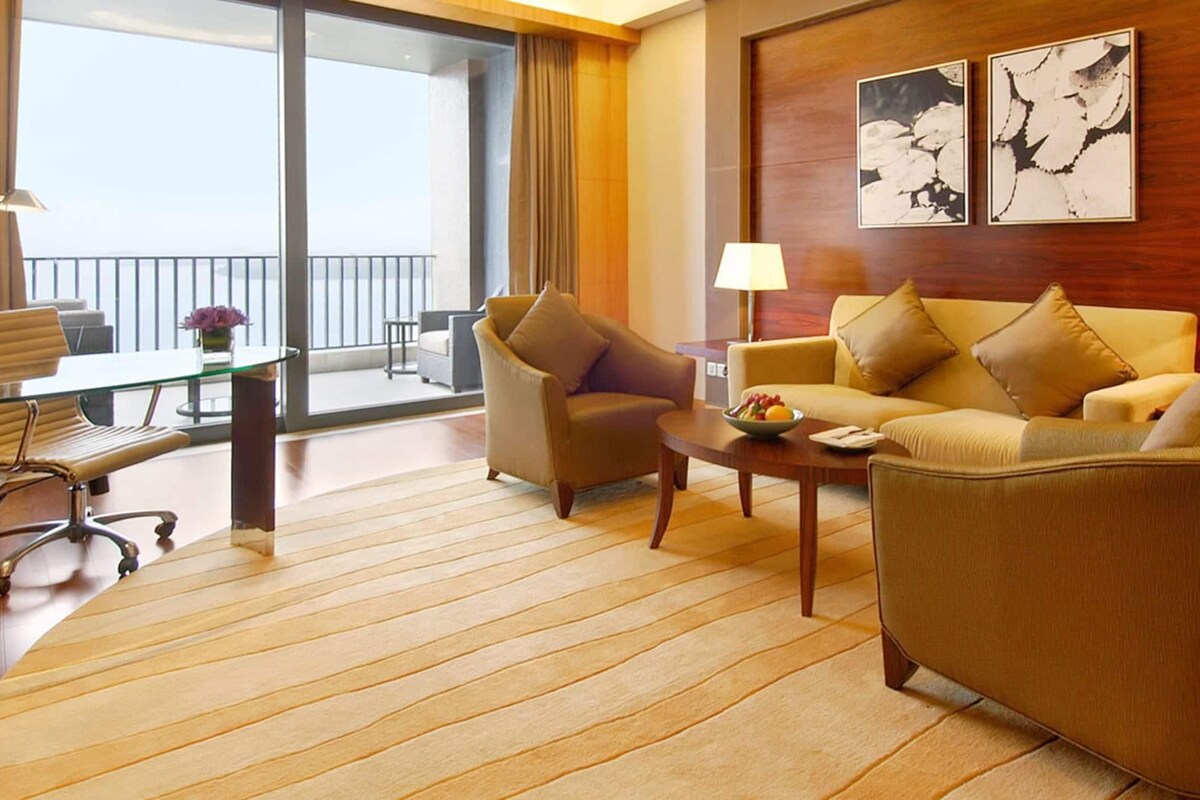 Habitación de hotel con sofá, sillón, mesa de centro y una mesa de comedor con sillas. Hay dos lámparas sobre la mesa, vistas al balcón y una obra de arte en la pared.