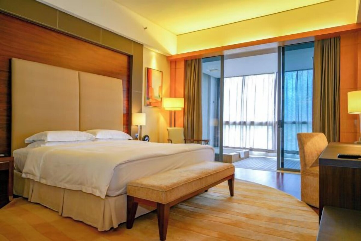 Habitación de hotel con una cama grande, una banca, un escritorio y vistas al exterior.