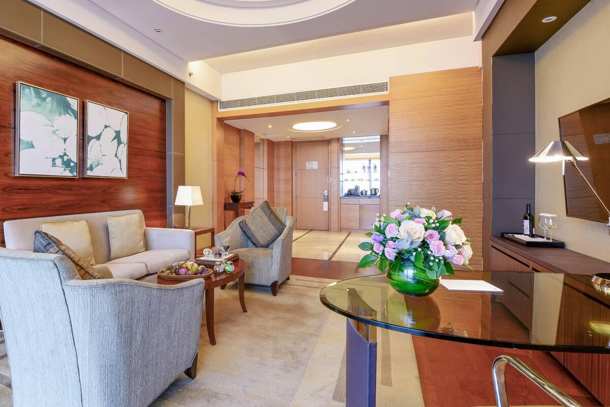 Un moderno vestíbulo de hotel con un sofá, sillones, una mesa de centro de cristal y un comedor con una lámpara y flores.