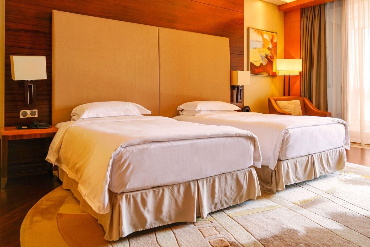 Habitación de hotel con dos camas, cabecera de madera, escritorio con lámpara, una silla y un amplio ventanal con cortinas.