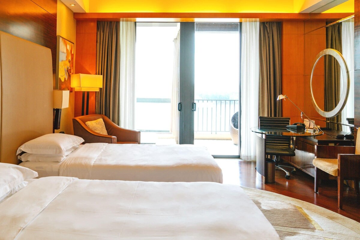 Habitación de hotel con dos camas, un escritorio y una silla, un sofá, un espejo grande y vistas al exterior.