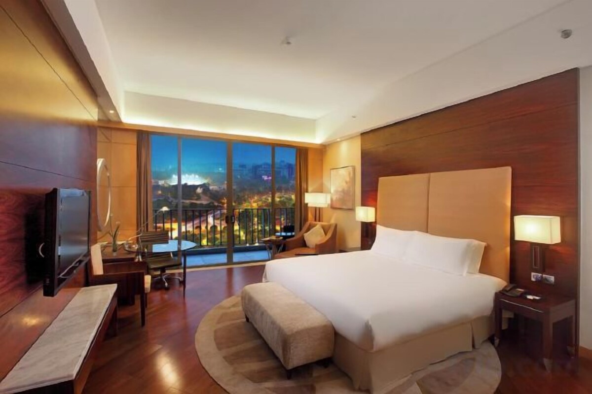 Habitación de hotel con una cama grande, un escritorio, una silla, un televisor y vistas a un paisaje urbano nocturno.