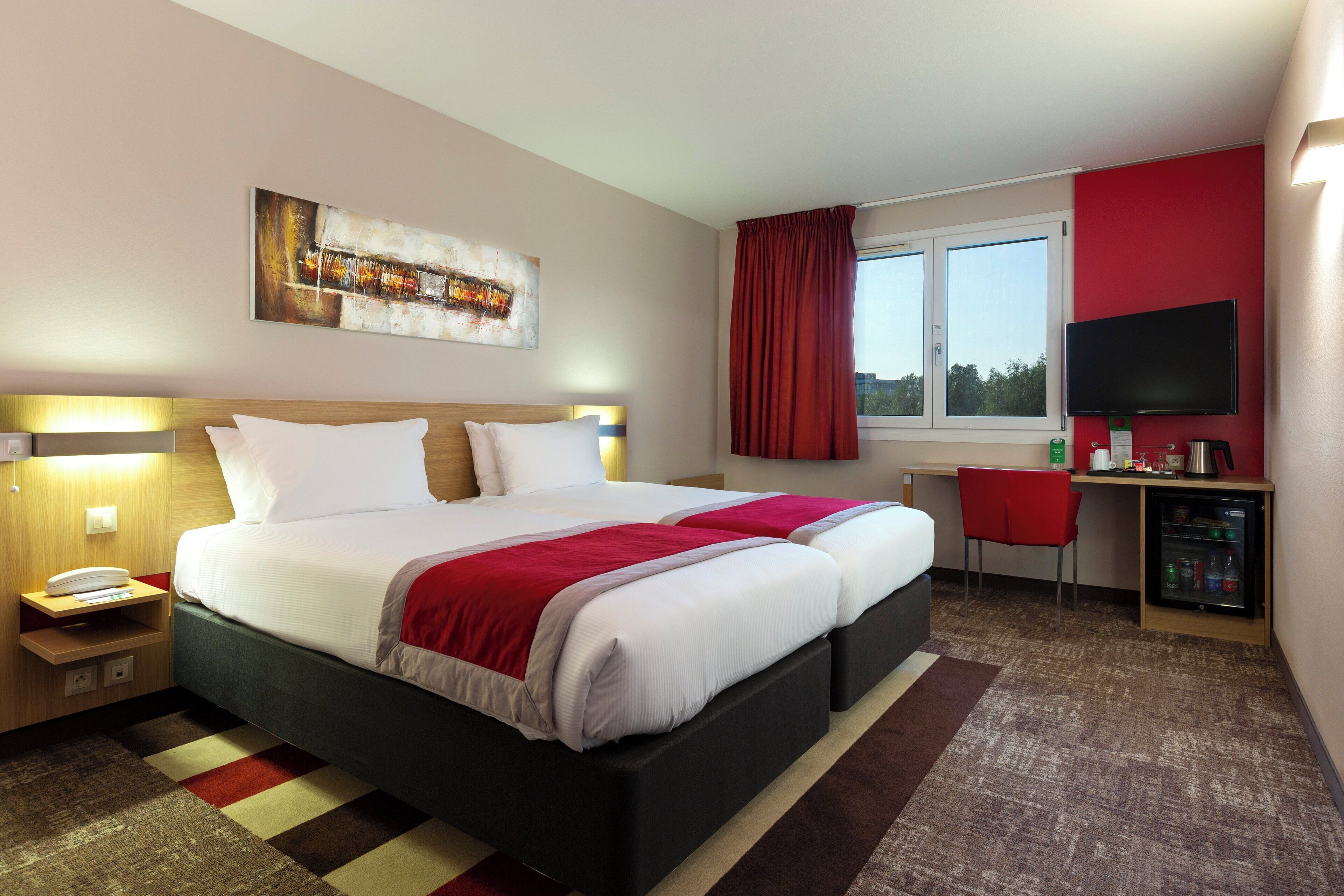 Een hotelkamer met een bed, nachtkastje, bureau, stoel, televisie en een raam met gordijnen.