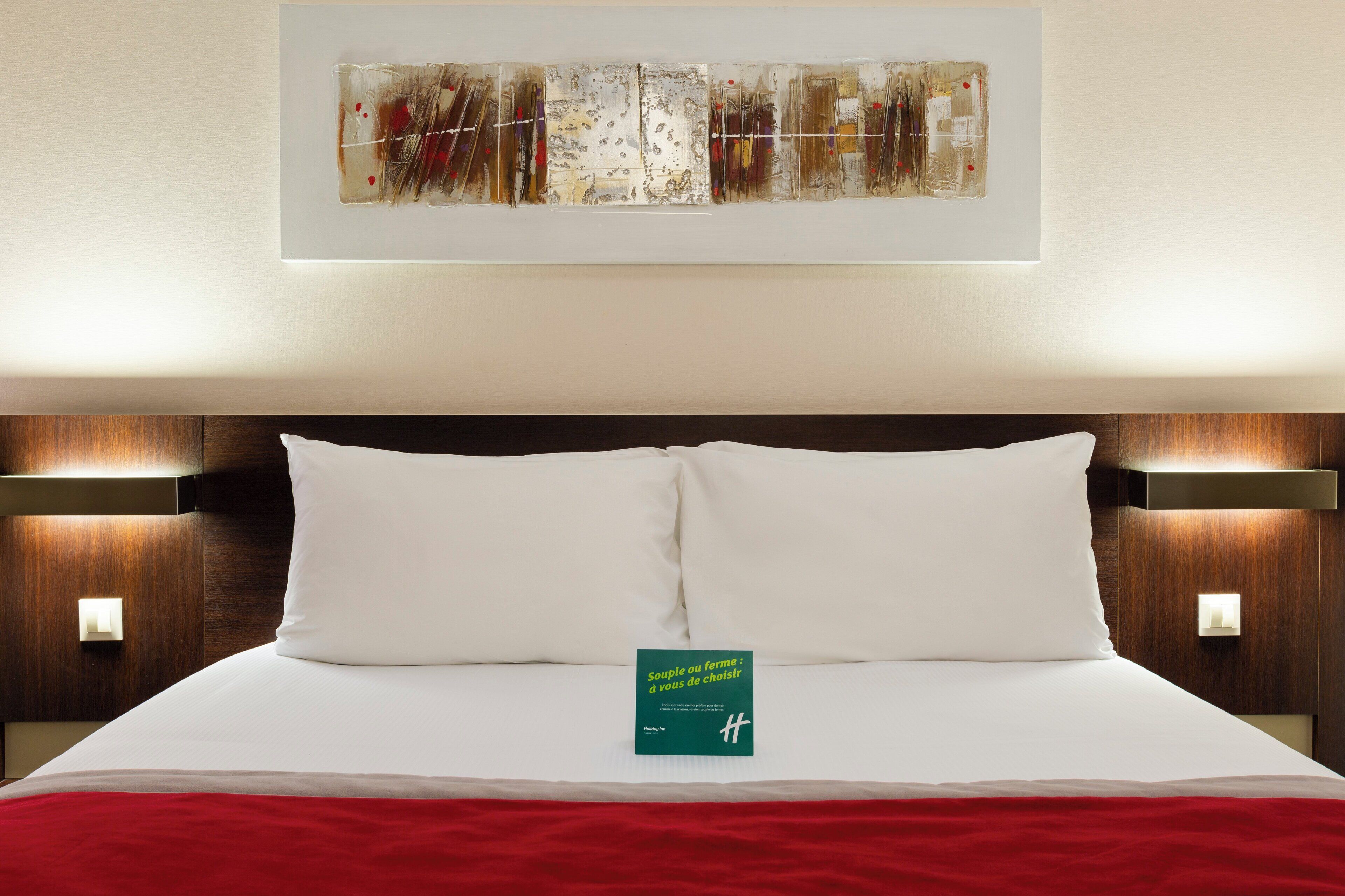 A hotel room with a neatly made bed, a framed abstract painting above it, and a green book on the bed.