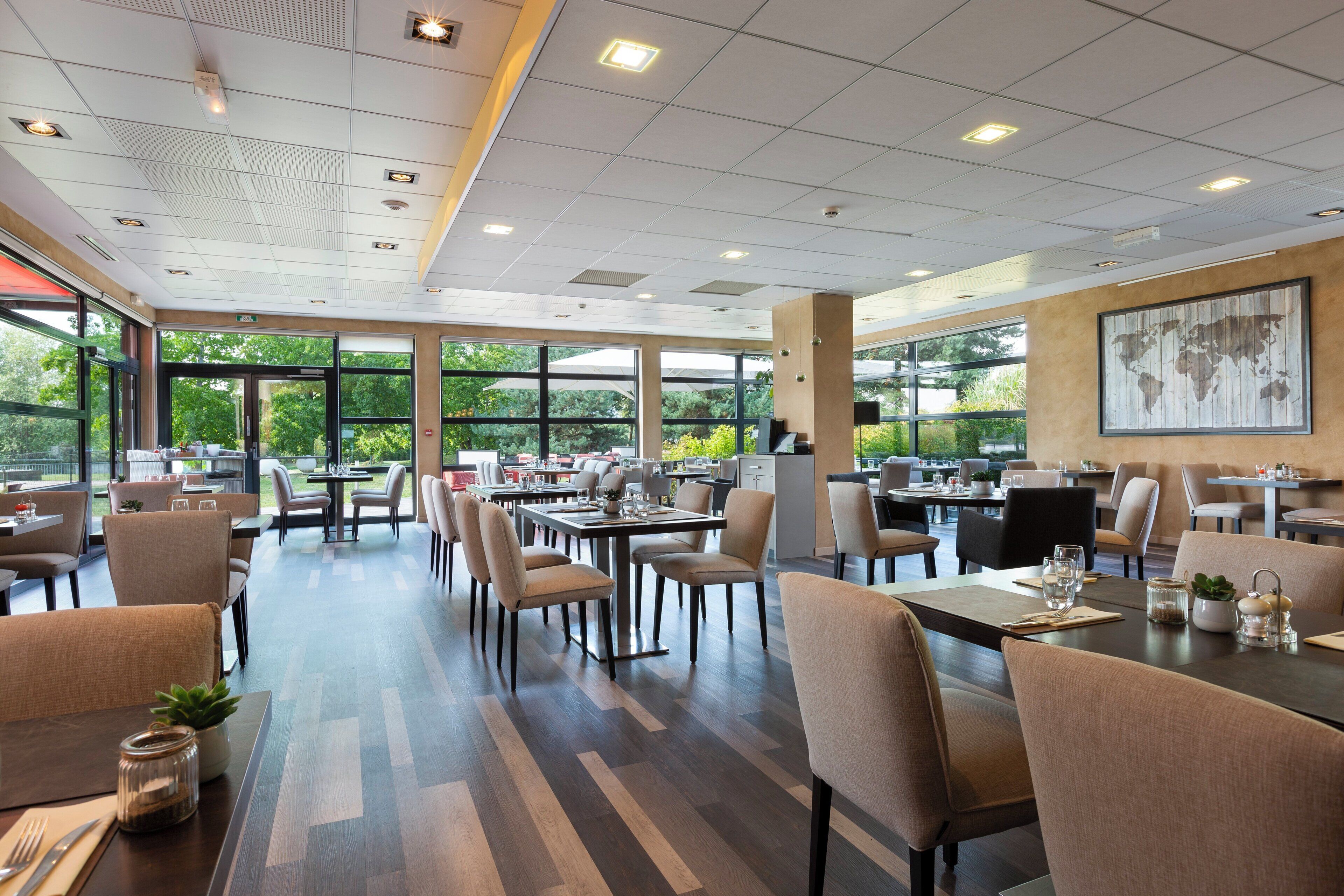 Un restaurant spacieux, doté de nombreuses tables et chaises, de grandes fenêtres offrant une vue sur la verdure et d’un plafond au design moderne.
