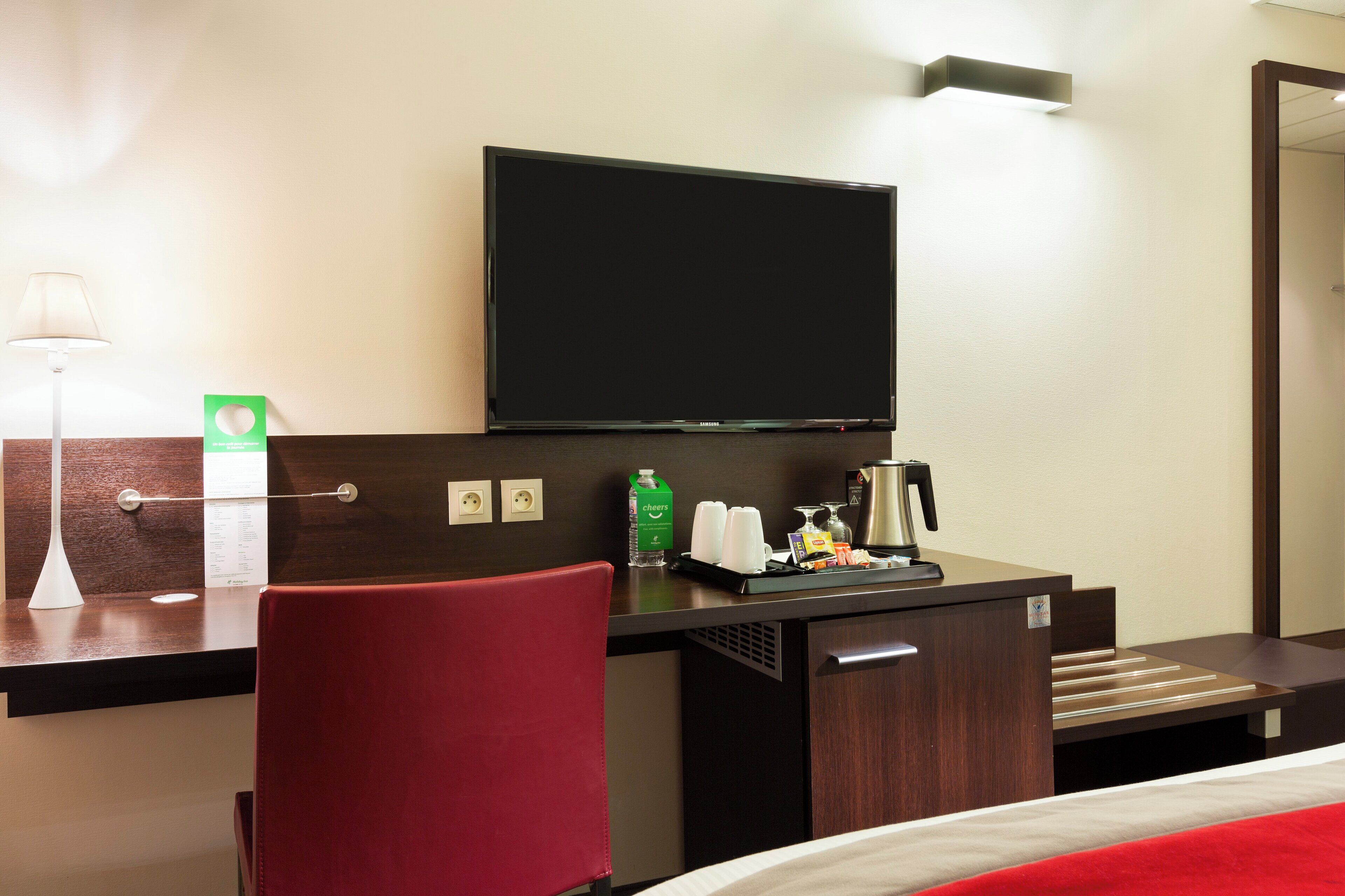 A hotel room with a bed, a desk with a flat-screen TV, a red chair, a coffee maker, and a bottle of water.