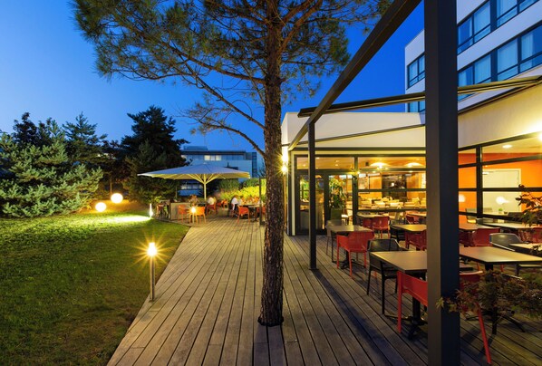 An outdoor dining area with tables and chairs, a wooden deck, and a building with large windows. - Holiday Inn Strasbourg - Nord by IHG (Schiltigheim)