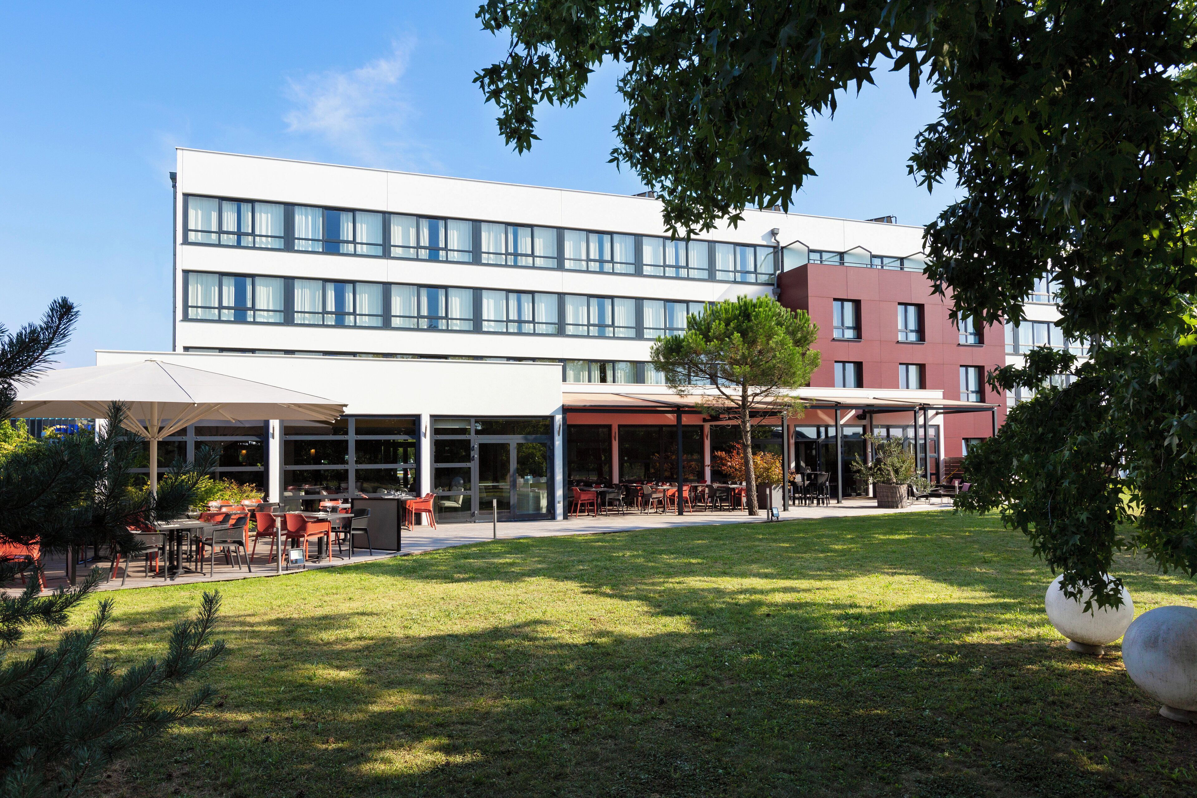 Un bâtiment moderne doté d’un espace extérieur aménagé, partiellement abrité, et d’une cour verdoyante.