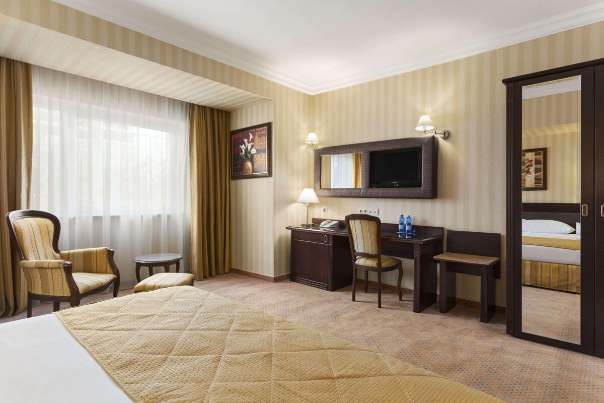 Een hotelkamer met een bed, een bureau, twee stoelen, een televisietoestel en een grote spiegel.