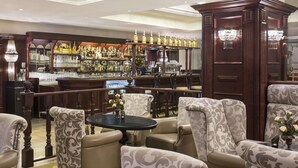 Uma área de bar com balcão de madeira, banquetas e um candelabro. Há cadeiras com estofamento estampado e uma mesa com um vaso de flores.