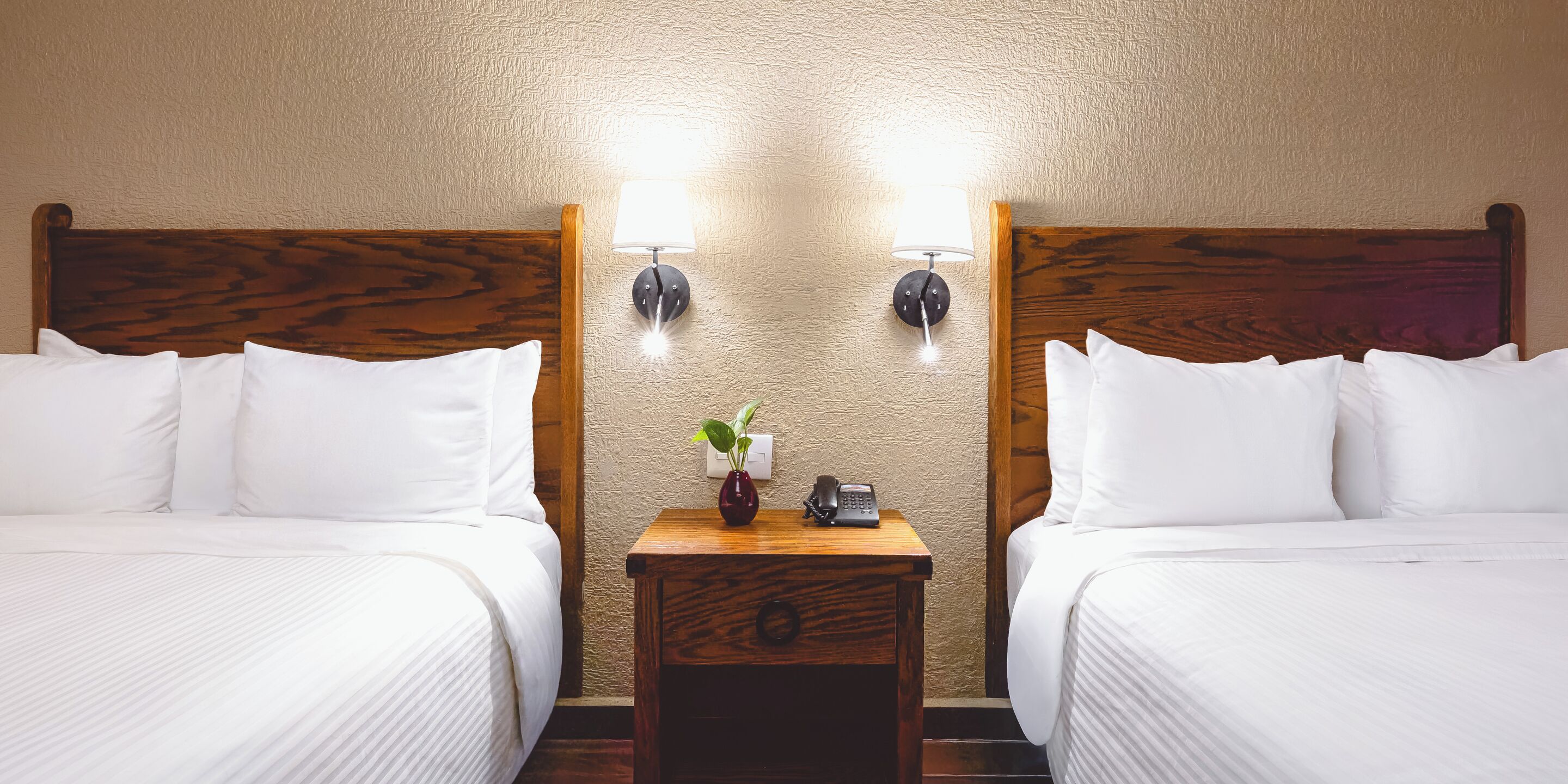 A hotel room with two beds, wooden headboards, white bedding, a nightstand with a lamp, a vase with a plant, and a phone.