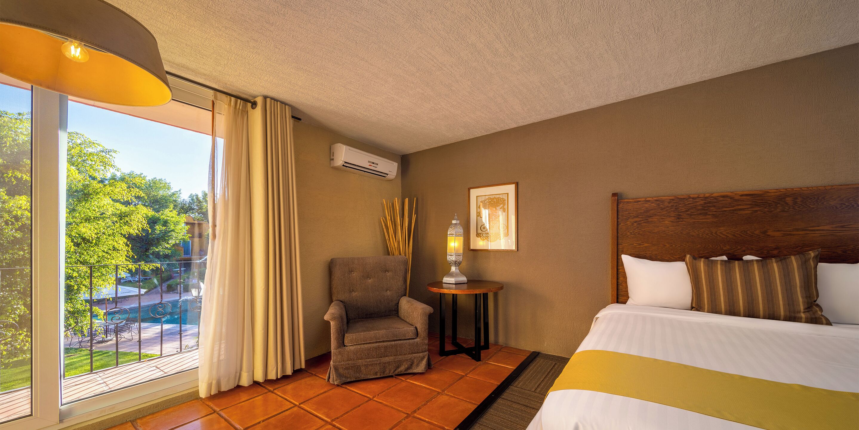 A hotel room with a bed, a chair, a small table with a lamp, and a window with curtains.