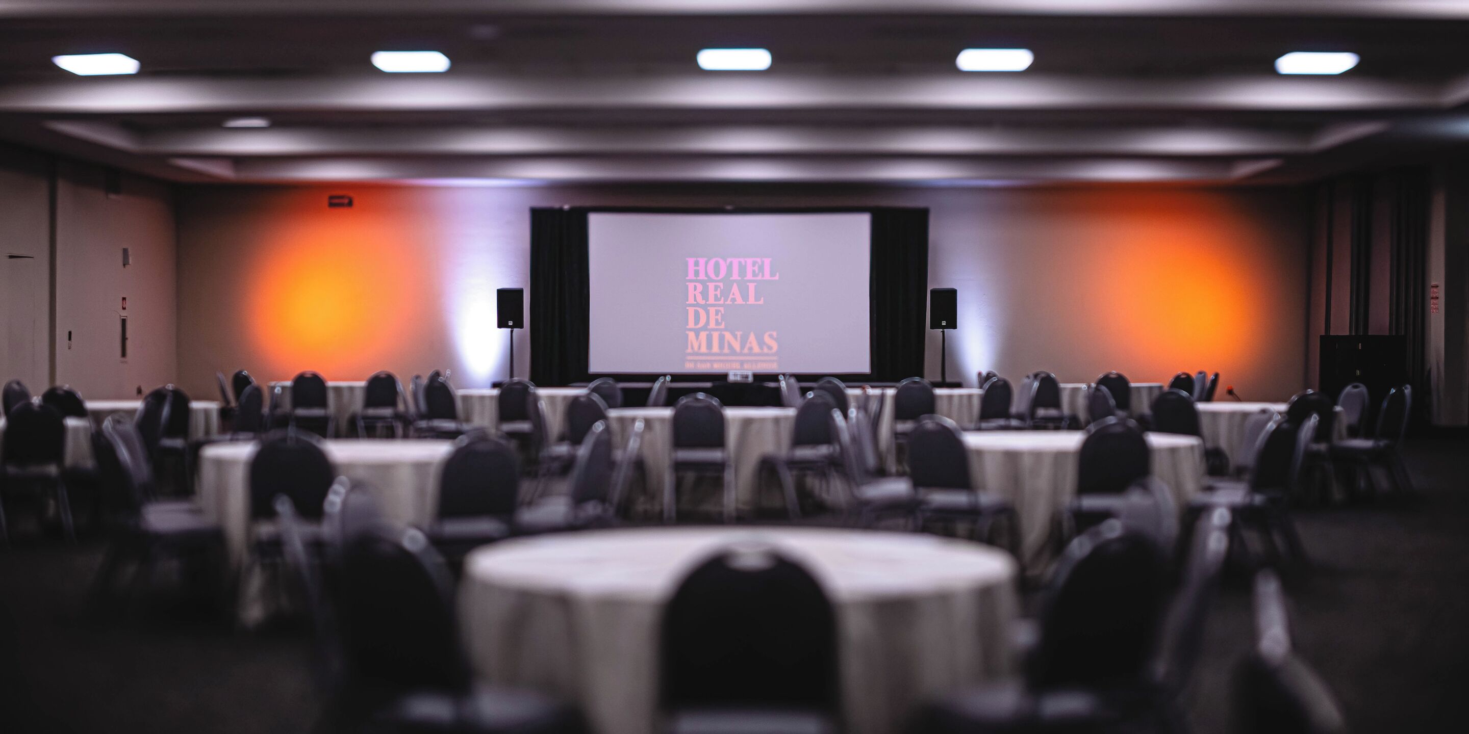 A hotel conference room with round tables and chairs, a large screen displaying 'HOTEL REAL DE MINAS', and speakers on either side.