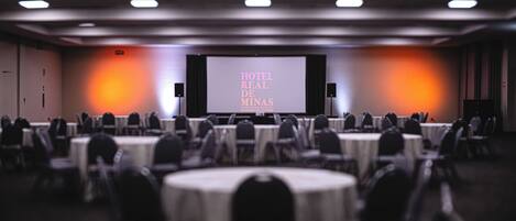 A hotel conference room with round tables and chairs, a large screen displaying 'HOTEL REAL DE MINAS', and speakers on either side.