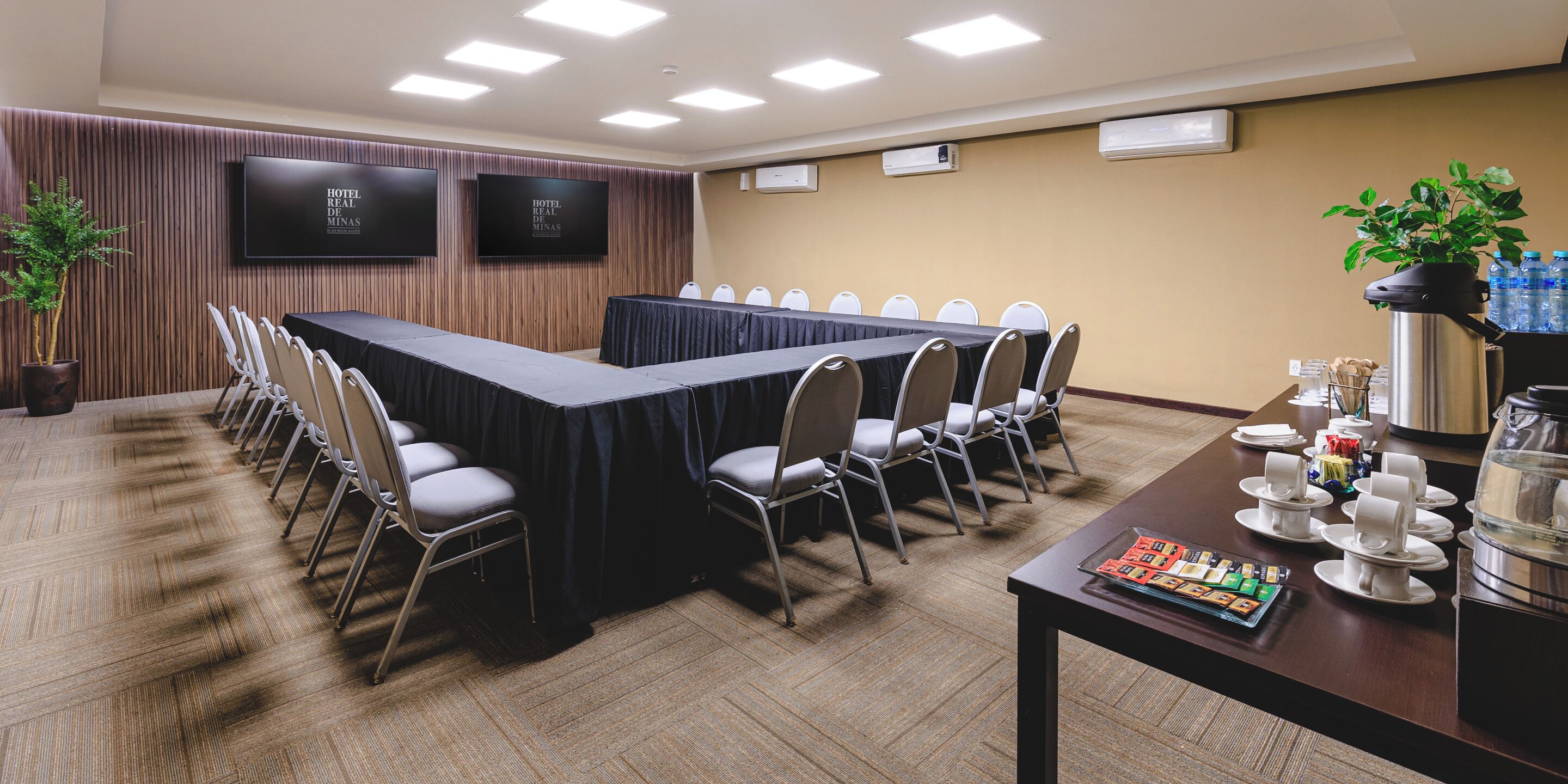 Una sala de conferencias con una mesa larga, sillas, una estación de café y dos televisores en la pared.