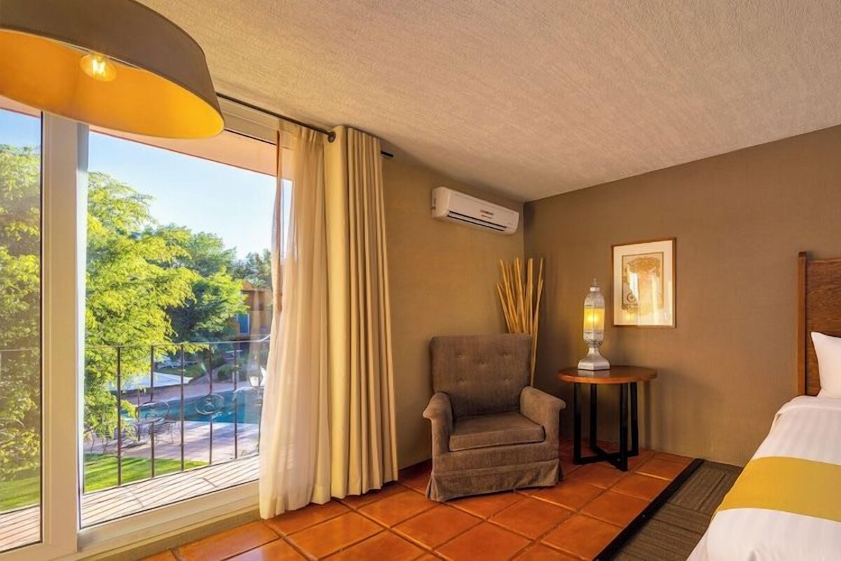 A hotel room with a bed, a chair, a small table with a lamp, an air conditioner, and a window with curtains.