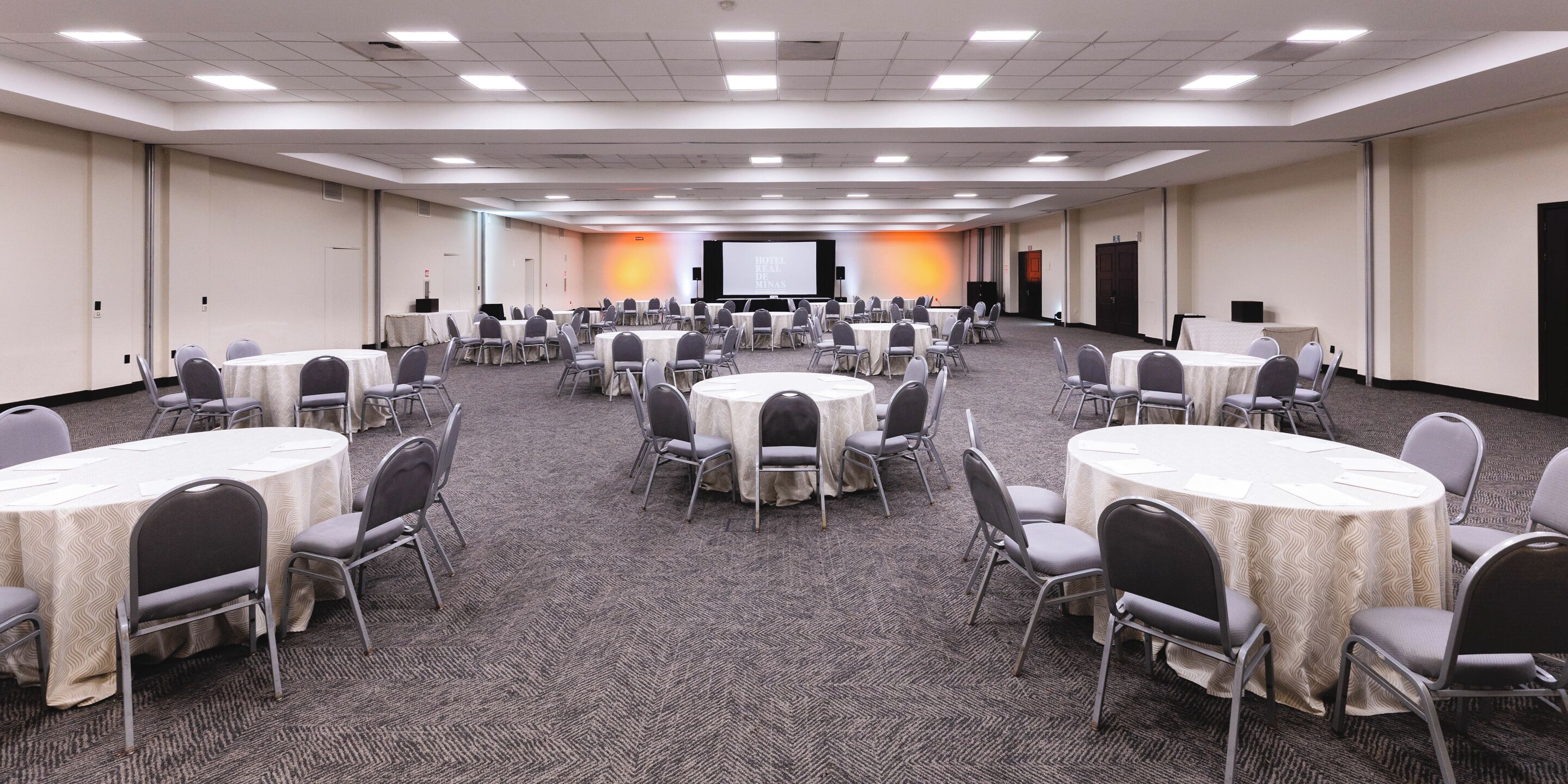A large conference room with multiple round tables and chairs, carpeted floor, and a projection screen at the front.