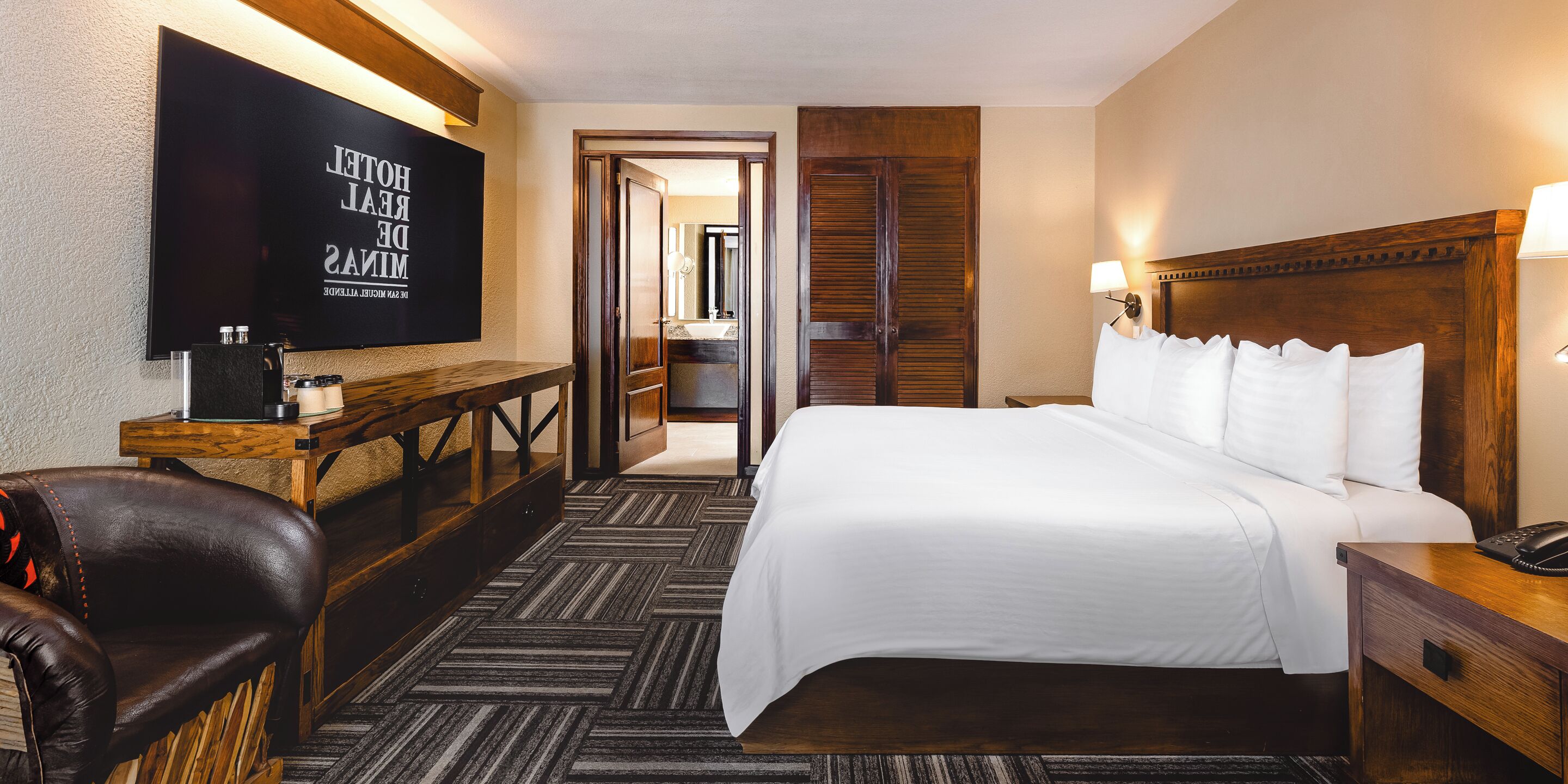 A hotel room with a bed, bedside table, lamp, TV, and a wooden bench.