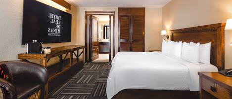 A hotel room with a bed, bedside table, lamp, TV, and a wooden bench.