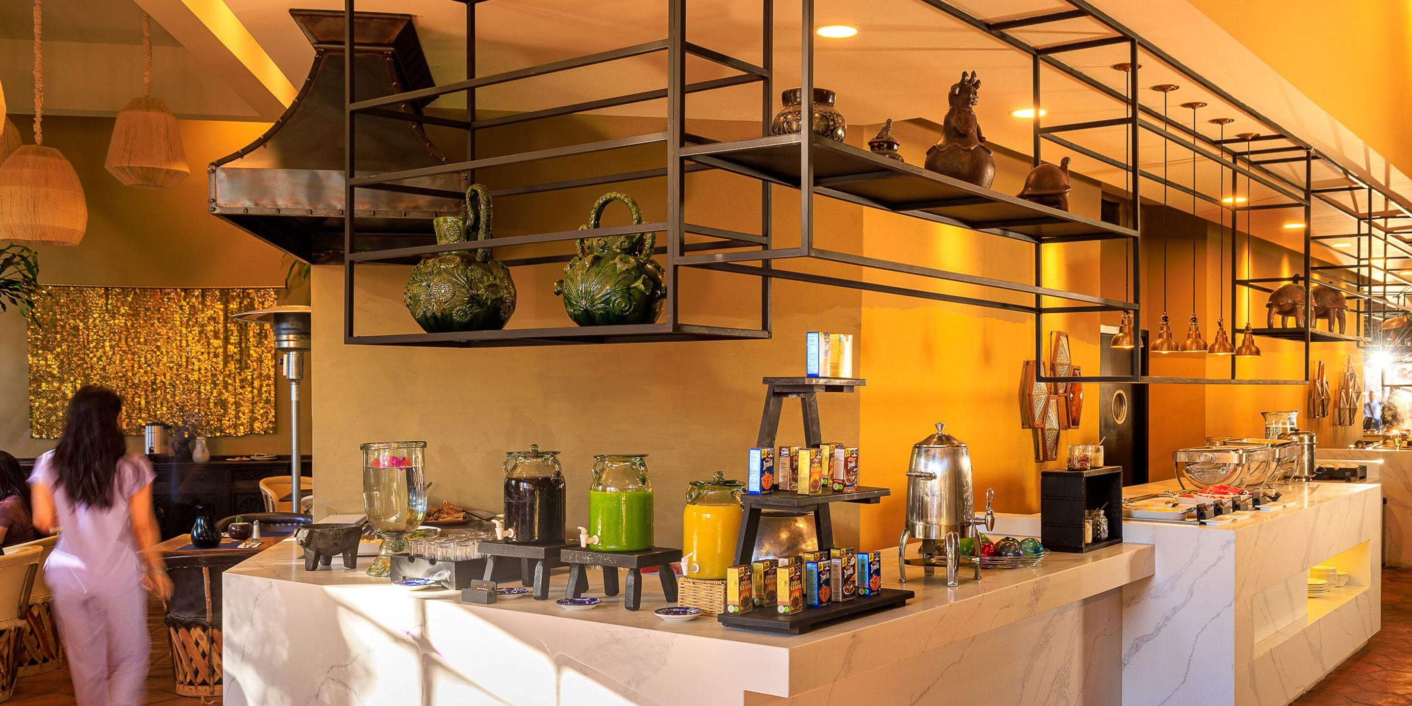 A breakfast bar with various food items and beverages, a coffee machine, and decorative vases on shelves.